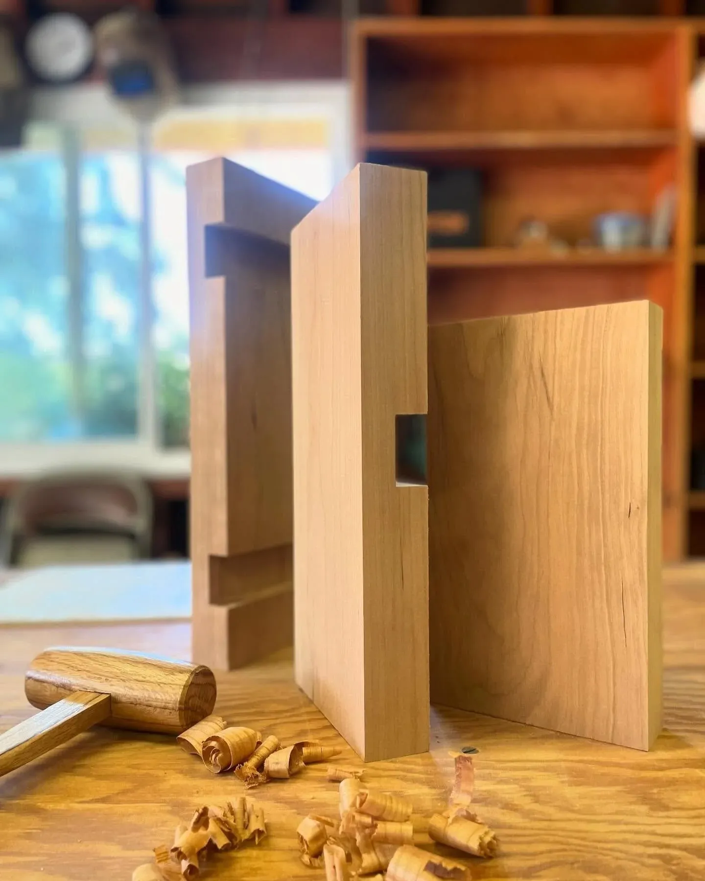 Two boards standing on a bench, one cut through with a rectangular mortise, a wooden mallet and hand-cut shavings beside them.