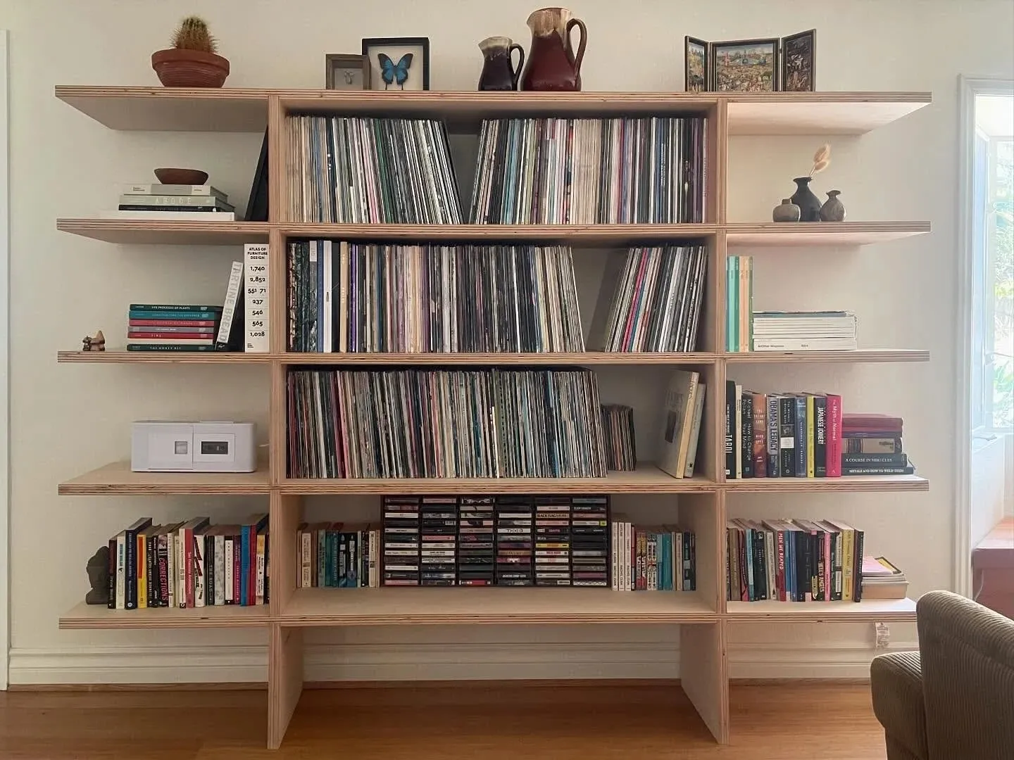 A full-height plywood shelving unit built into a room, filled with records, books and cassettes, with a window at the right.