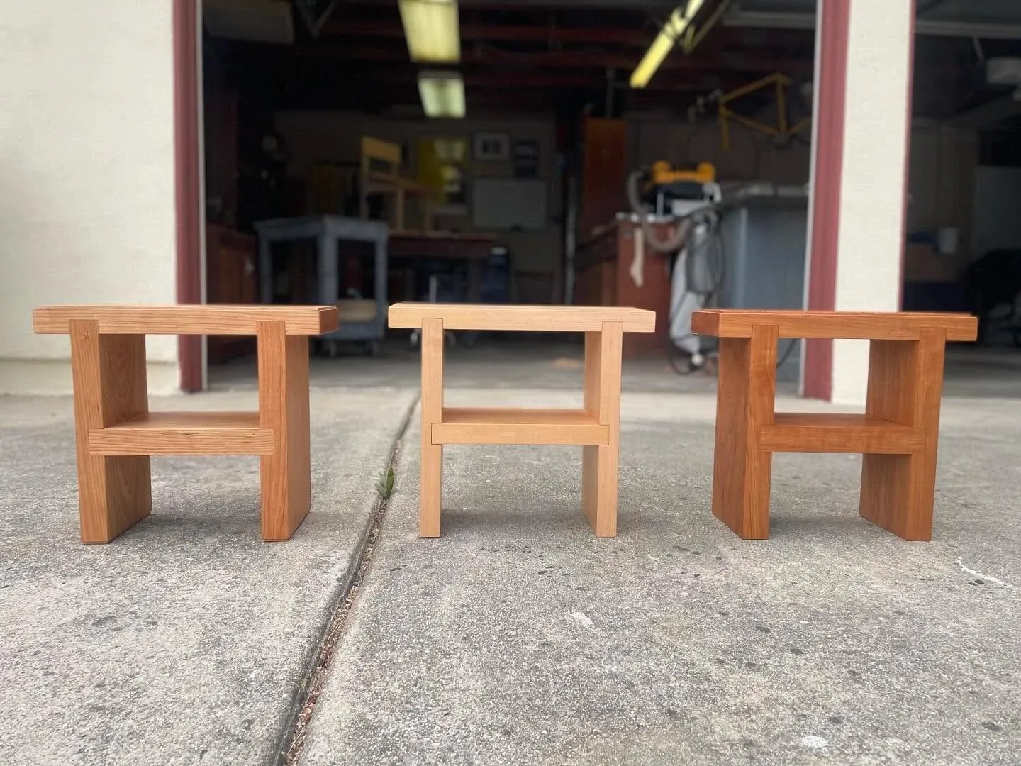Three matching low wooden stools standing in a row on a concrete apron outside the open workshop door.