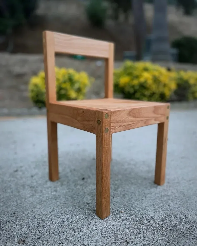 A small wooden chair seen from three-quarters on a concrete driveway, with round brass pegs showing at the seat corners.